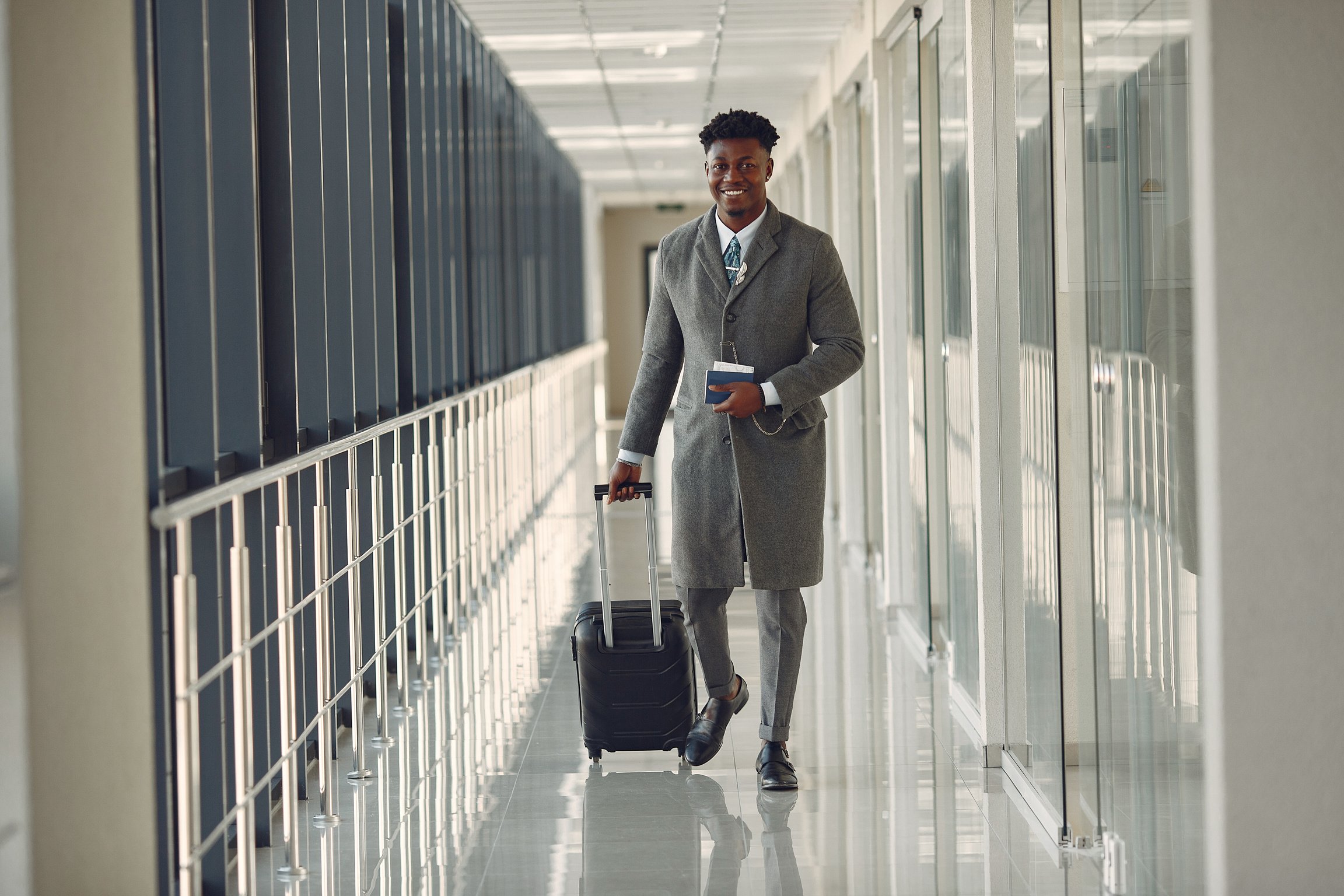 Stylish man with suitcase and passport walking along airport corridor